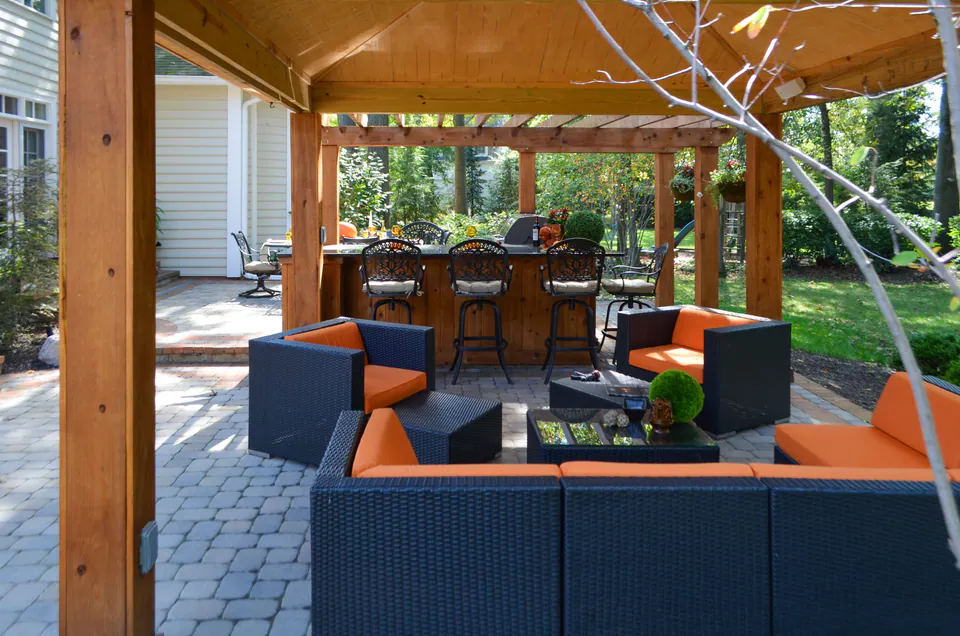 Interior of covered pavilion with orange lounge seating, wood ceiling, paver floor, string lights, and open backyard view.